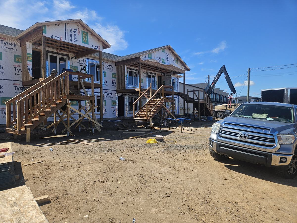 Two-storey homes in framing stage with Tyvek house wrap and wooden stairs installed on sandy lot.