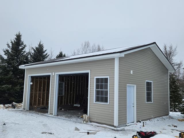 Newly framed garage with vinyl siding, white trim, and garage doors during winter construction.