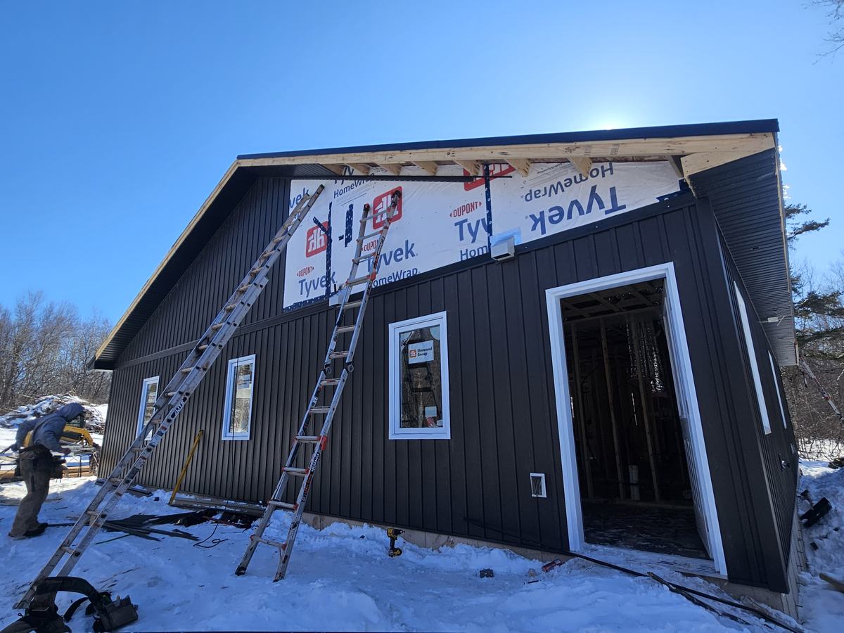 Black metal siding being installed on a residential structure with white trim and weatherproofing wrapping visible.